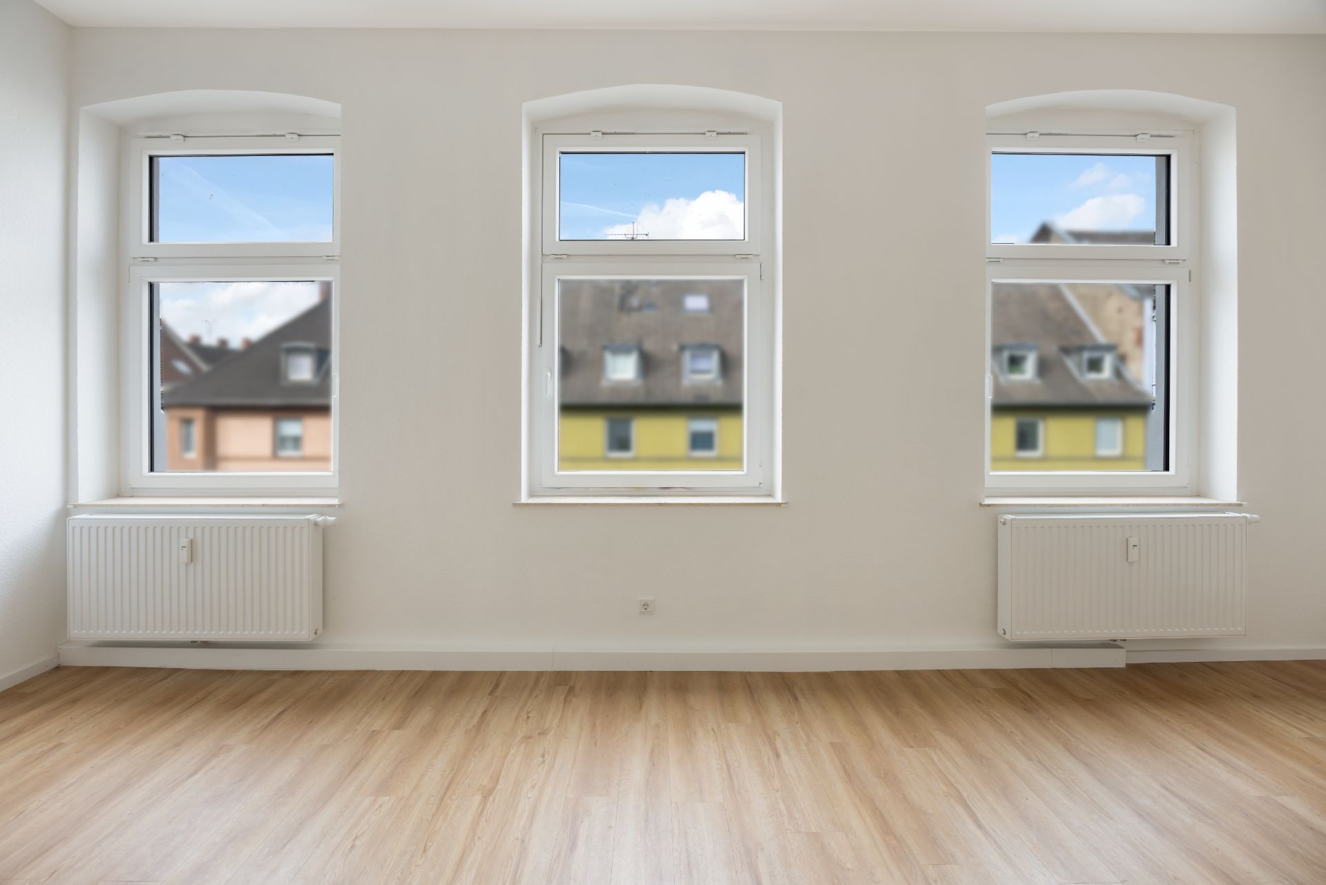 Heller Raum mit Holzfußboden, drei Fenstern und zwei Heizkörpern unter den Fenstern.
