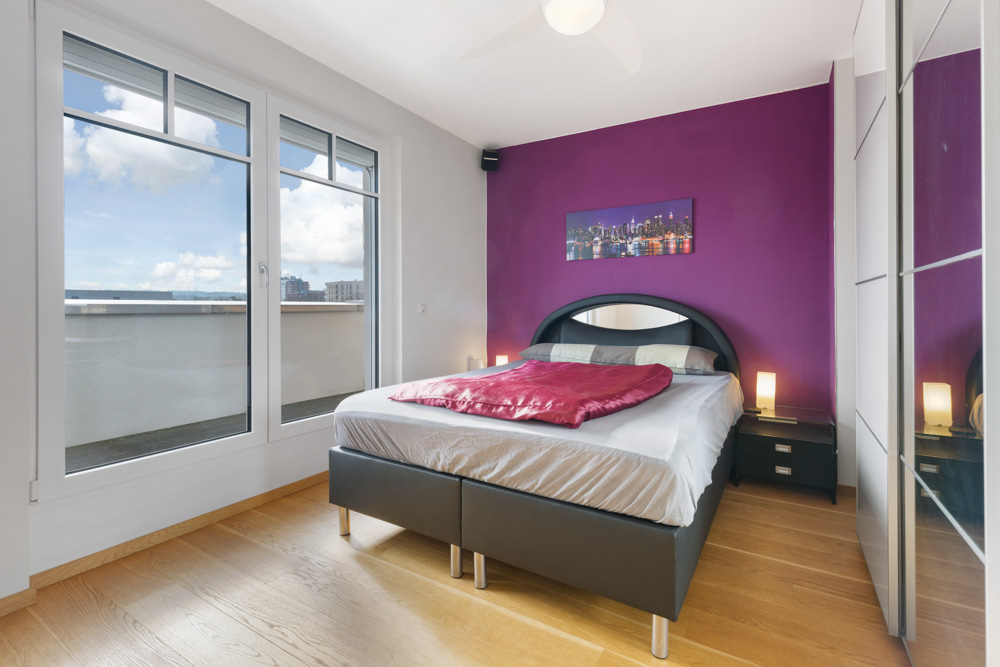 Modernes Schlafzimmer mit großem Bett, violetter Wand, großen Fenstern und Holzfußboden.