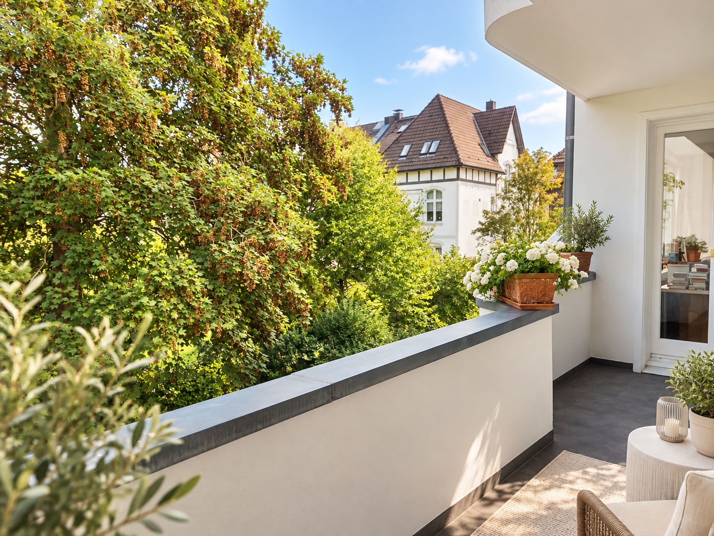 Heller Balkon mit Pflanzen, Blick auf Bäume und Nachbarhaus mit Satteldach.