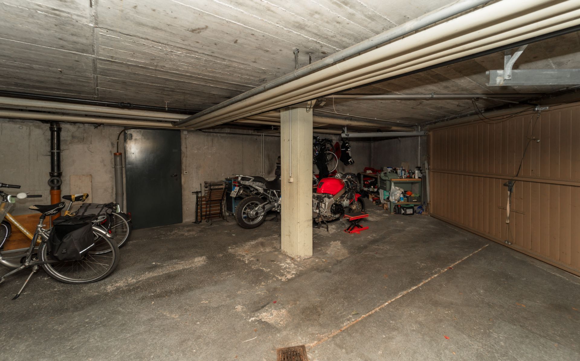 Garage mit Fahrrädern, einem Motorrad, Werkbank und Betonwänden.