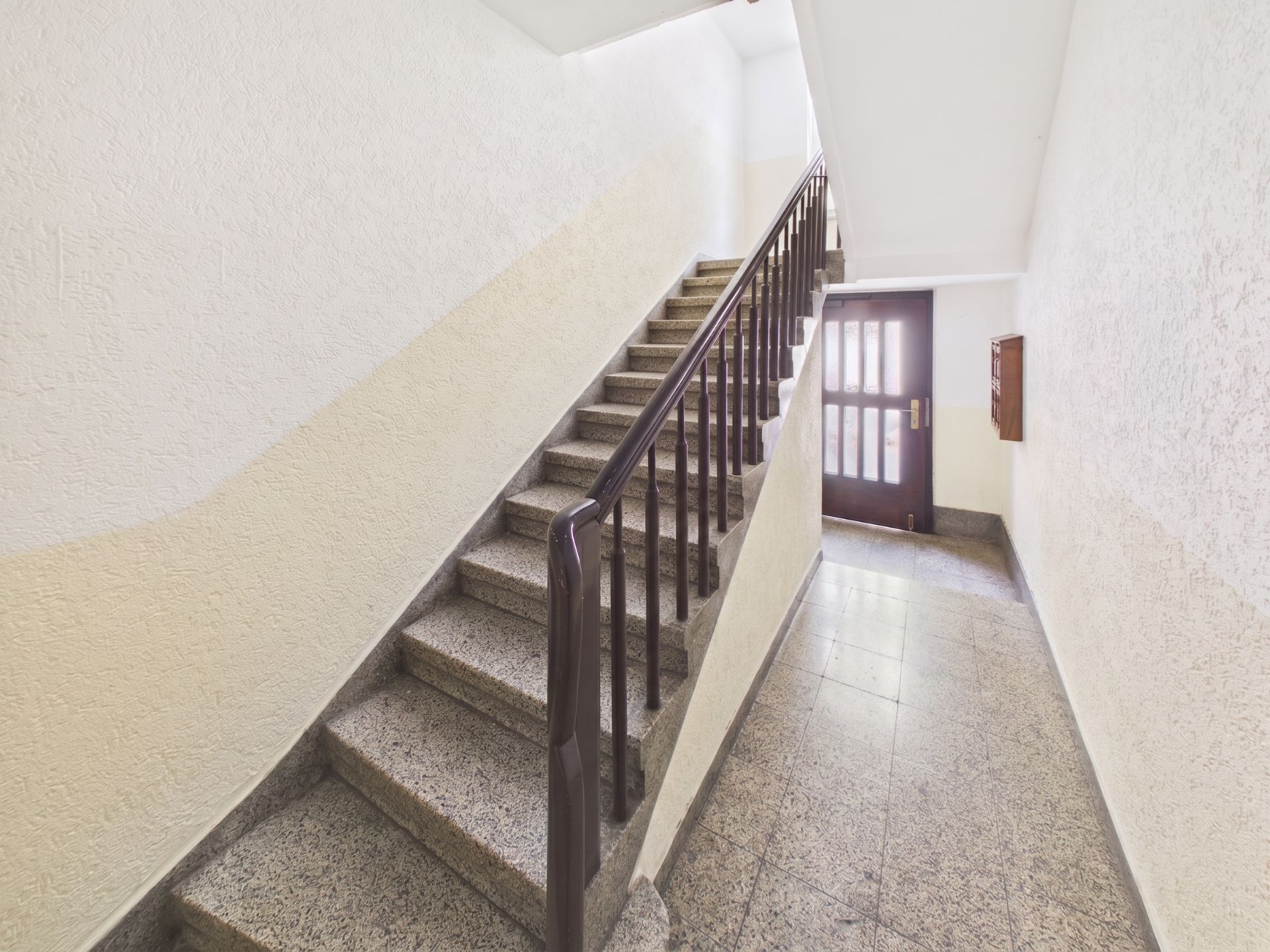 Helles Treppenhaus mit Steintreppe und Handlauf, am Ende eine Glastür.