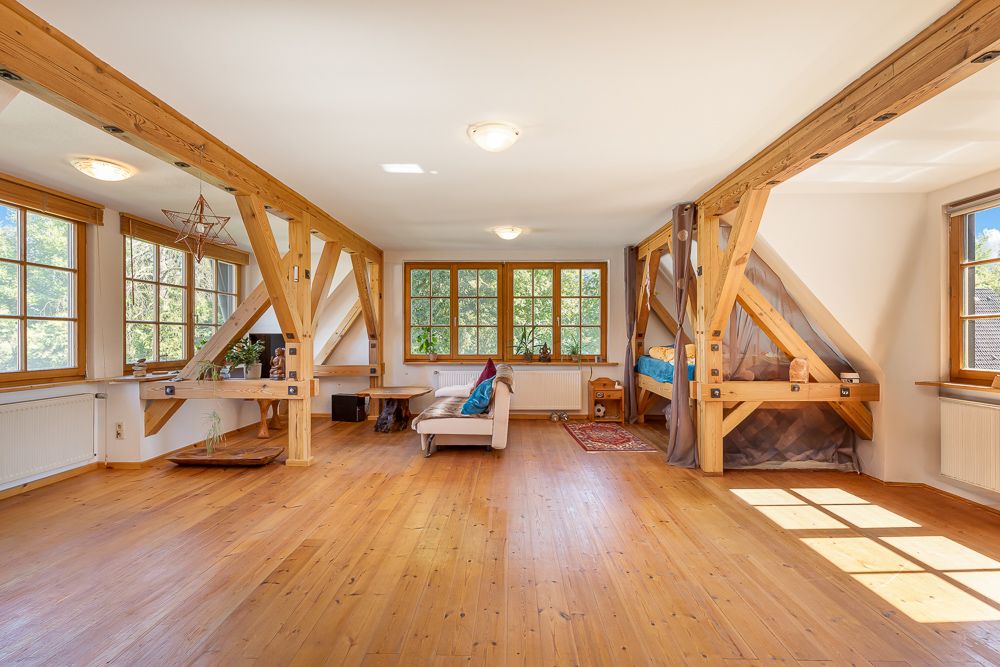 Heller Raum mit Holzboden, sichtbaren Balken, Sofa, Schreibtisch und einem Bett unter der Schräge.