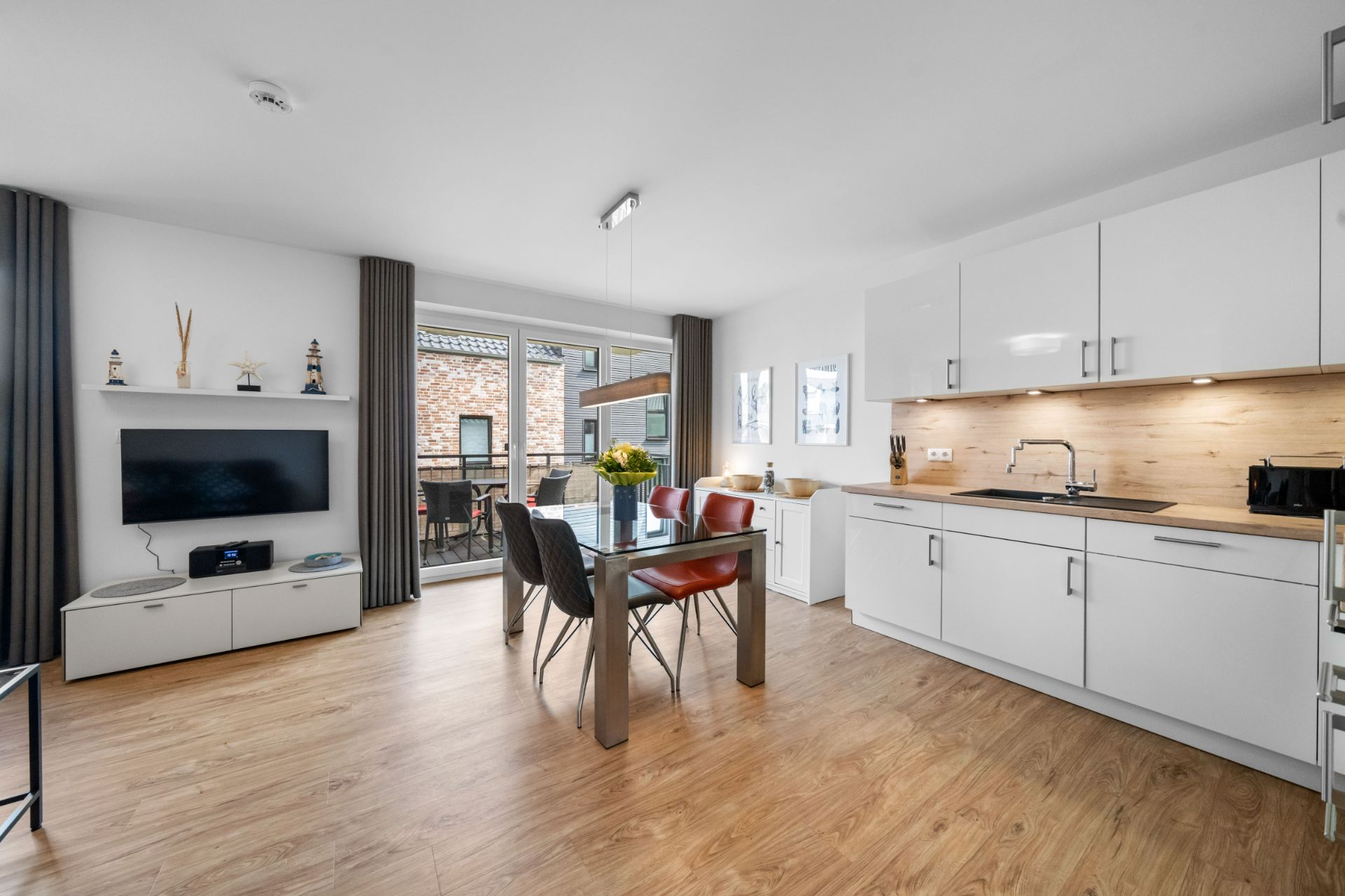 Helles Wohnzimmer mit offener Küche, Esstisch, Fernseher und Zugang zum Balkon.