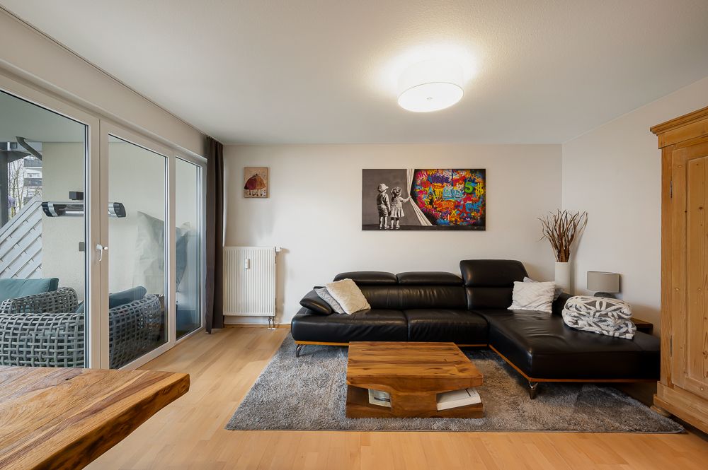 Wohnzimmer mit schwarzem Ecksofa, Holztisch, Teppich und Wandbildern. Zugang zum Balkon mit Sitzmöbeln.