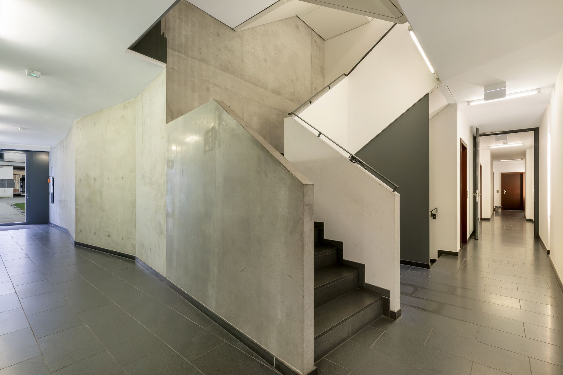 Heller Flur mit moderner Betontreppe, Türen und Fliesenboden in einem Wohngebäude.