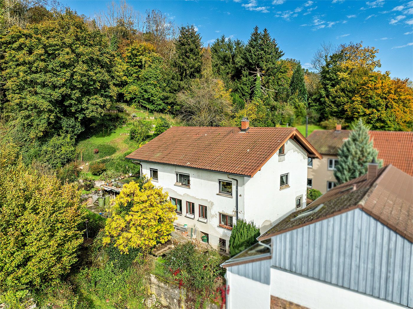 Weißes Wohnhaus mit rotem Dach, umgeben von Bäumen und Garten, unter blauem Himmel.