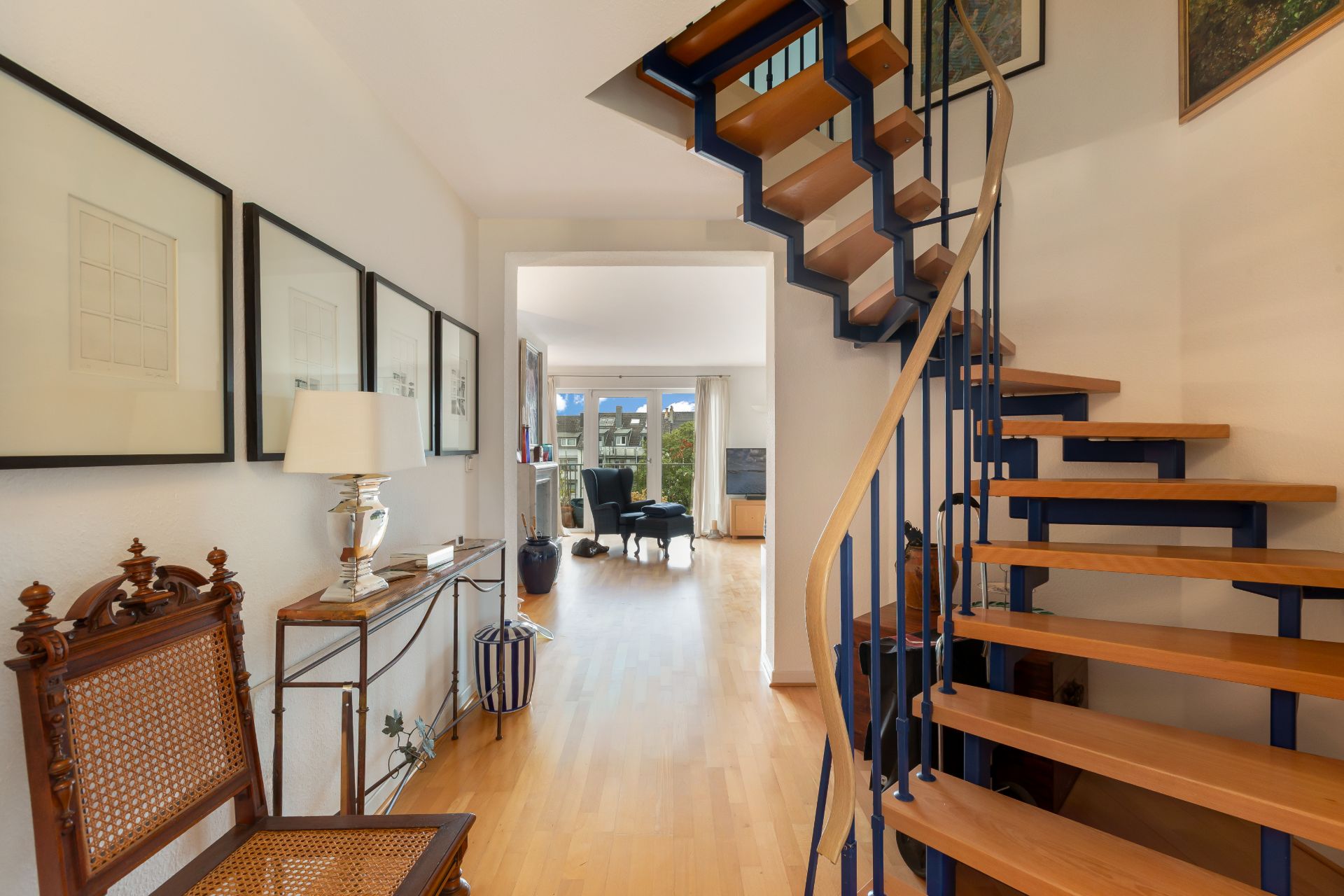 Heller Flur mit Holztreppe, Holzboden, Stuhl und Wandbildern, Blick ins Wohnzimmer.
