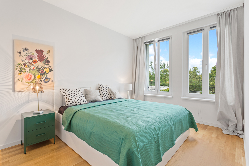 Helles Schlafzimmer mit großem Bett, grünem Nachttisch, Blumenbild und drei Fenstern.