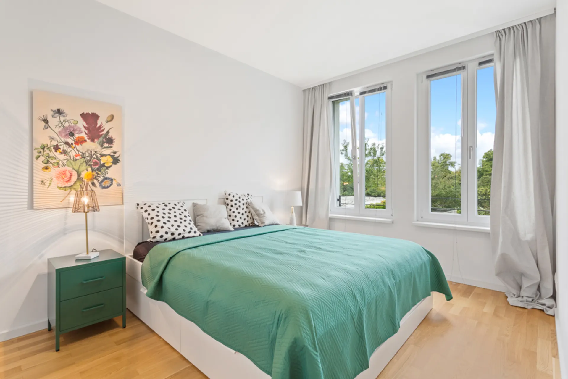 Helles Schlafzimmer mit großem Bett, grünem Nachttisch, Blumenbild und drei Fenstern.
