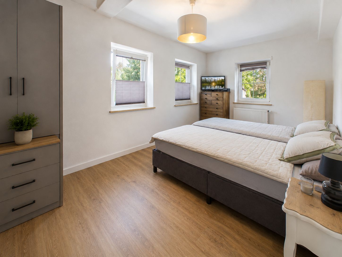 Helles Schlafzimmer mit Doppelbett, Kleiderschrank, Kommode und drei Fenstern.