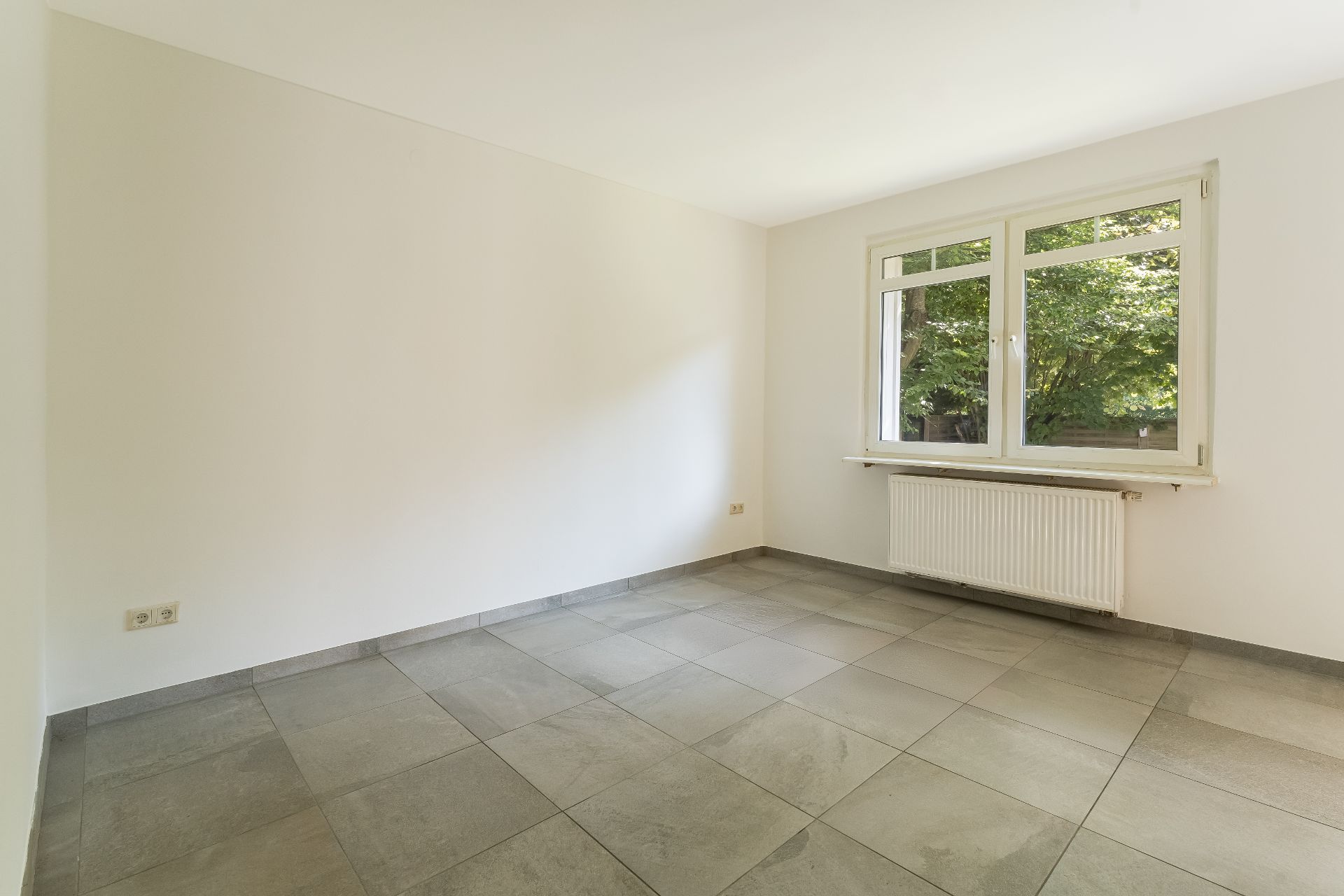 Leerer Raum mit Fliesenboden, weißer Wand und großem Fenster mit Blick ins Grüne.