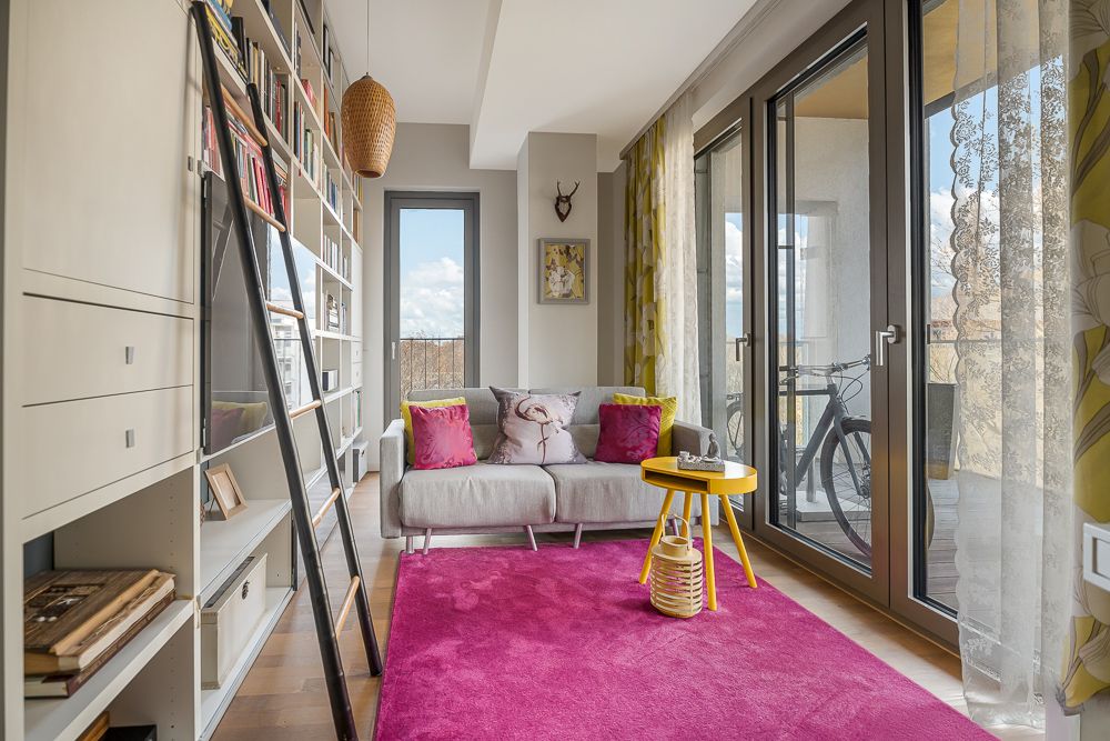 Helles Wohnzimmer mit Bücherregal, grauem Sofa, gelbem Tisch und pinkem Teppich vor großen Fenstern.