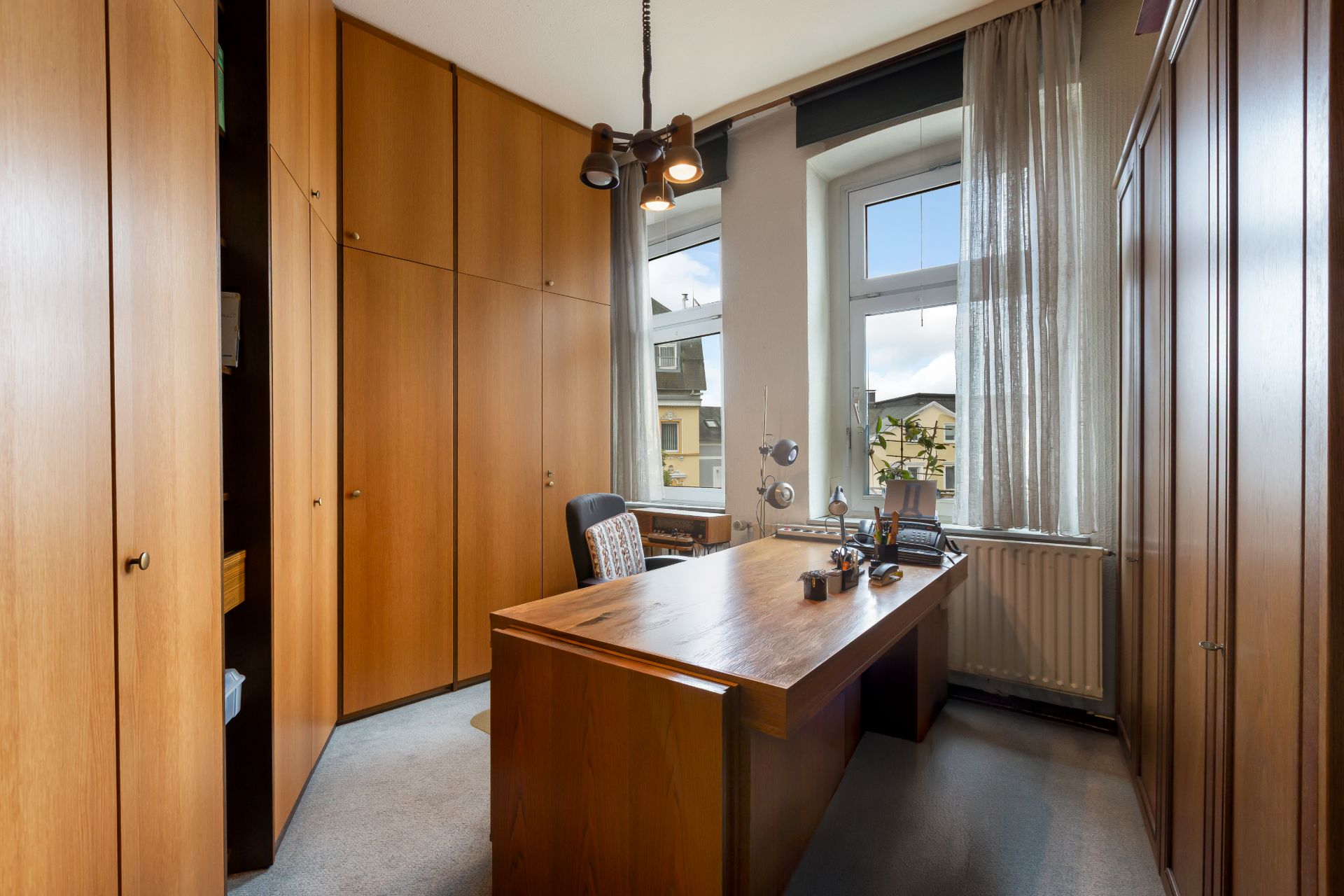 Büro mit Holzschreibtisch, Schränken, Stuhl und großem Fenster mit Vorhängen.