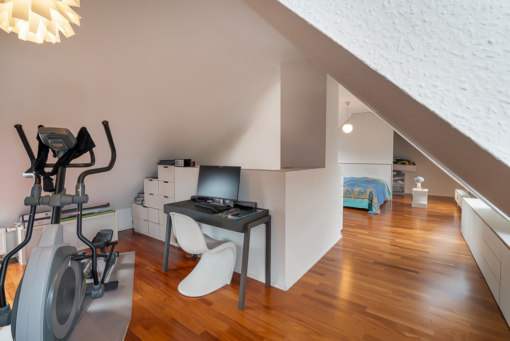 Helles Dachzimmer mit Fitnessgerät, Schreibtisch, Computer und Bett, Parkettboden.