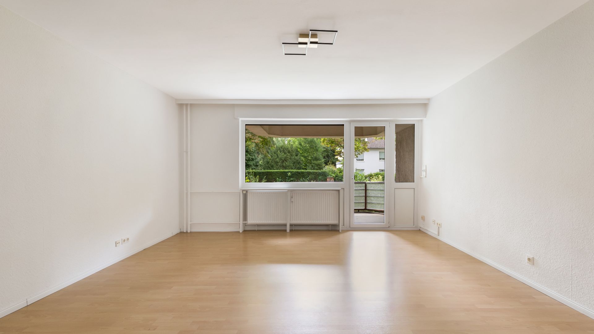 Leerer Raum mit Parkettboden, Fenster und Tür zu einem Balkon, Blick ins Grüne.