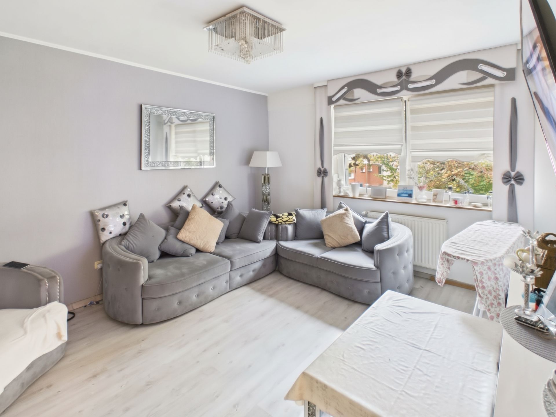Helles Wohnzimmer mit grauem Ecksofa, vielen Kissen, Spiegel und großen Fenstern mit Dekoration.