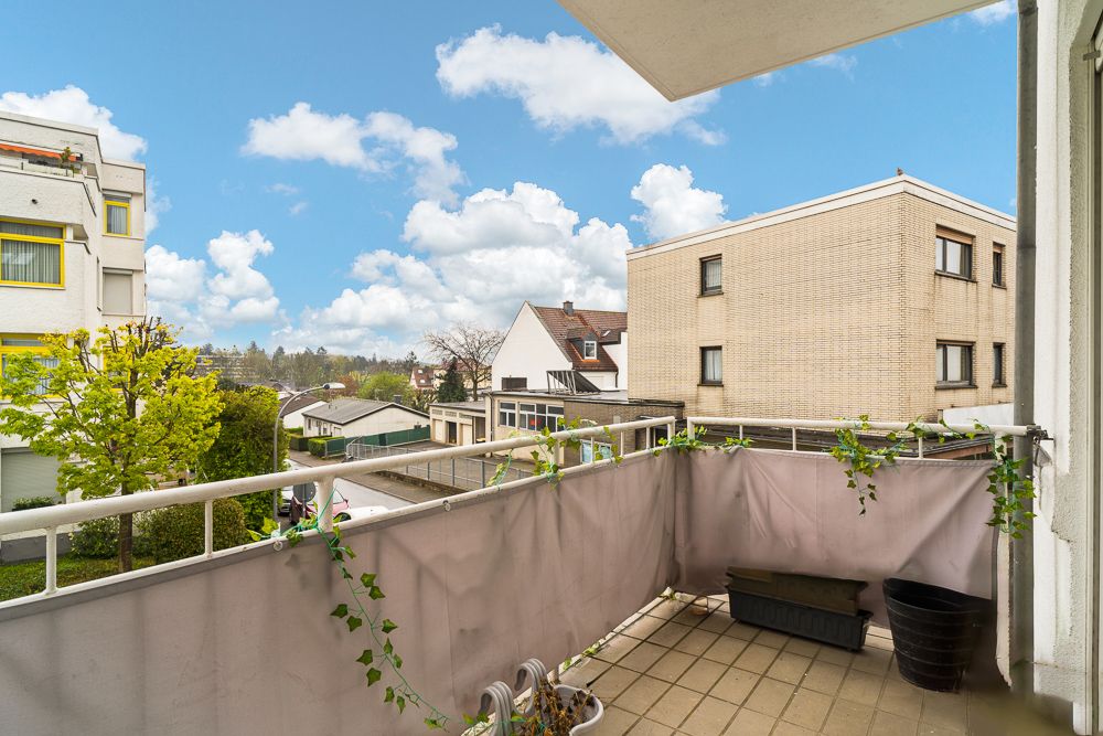Balkon mit Sichtschutz und Pflanzen, Blick auf Wohnhäuser und blauen Himmel mit Wolken.
