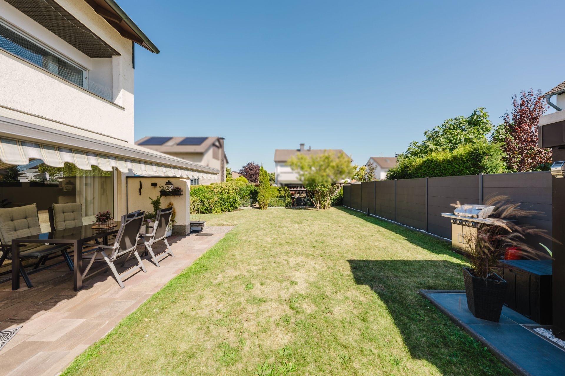 Garten mit Rasen, Terrasse mit Sitzgruppe und Grill, umgeben von Zaun und Nachbarhäusern.