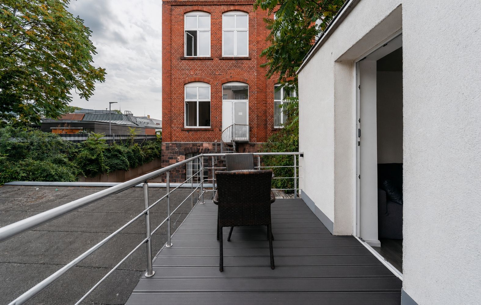 Balkon mit Stuhl und Tisch, Blick auf ein rotes Backsteingebäude und Grünflächen.