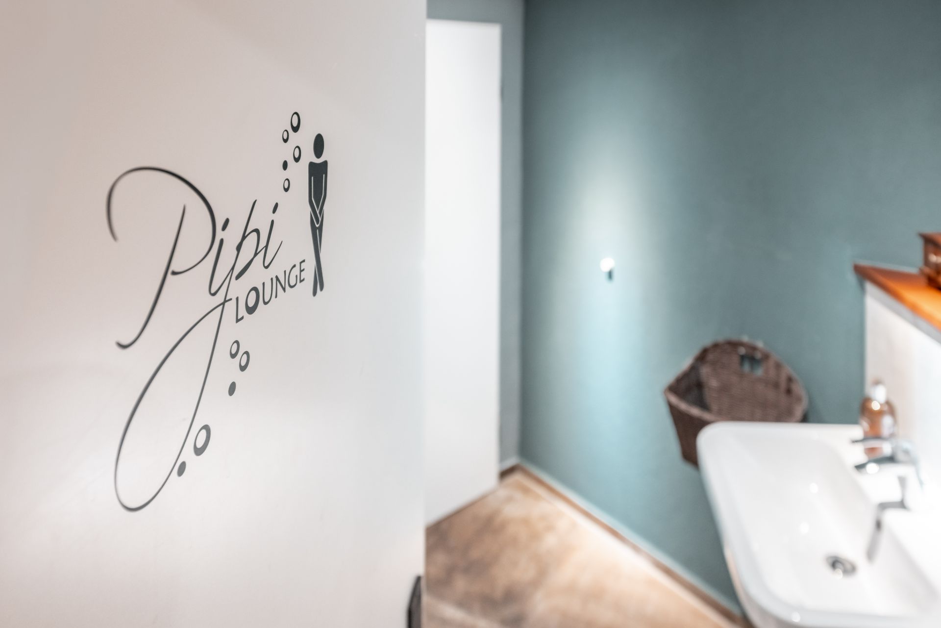 Offene Badezimmertür mit Aufschrift 'Pipi Lounge', Waschbecken und Korb an der Wand sichtbar.