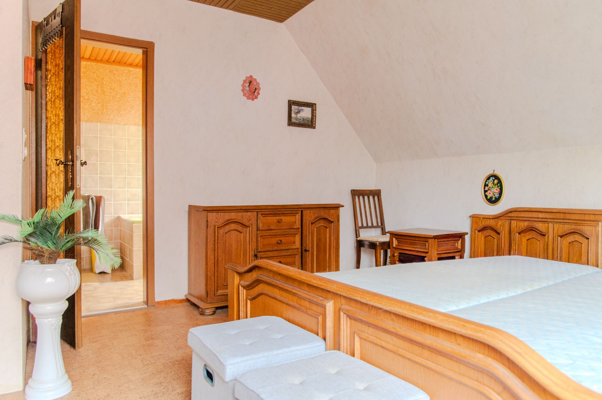 Helles Schlafzimmer mit Holzmöbeln, Doppelbett, Kommode, Stuhl, Topfpflanze und Blick ins Badezimmer.
