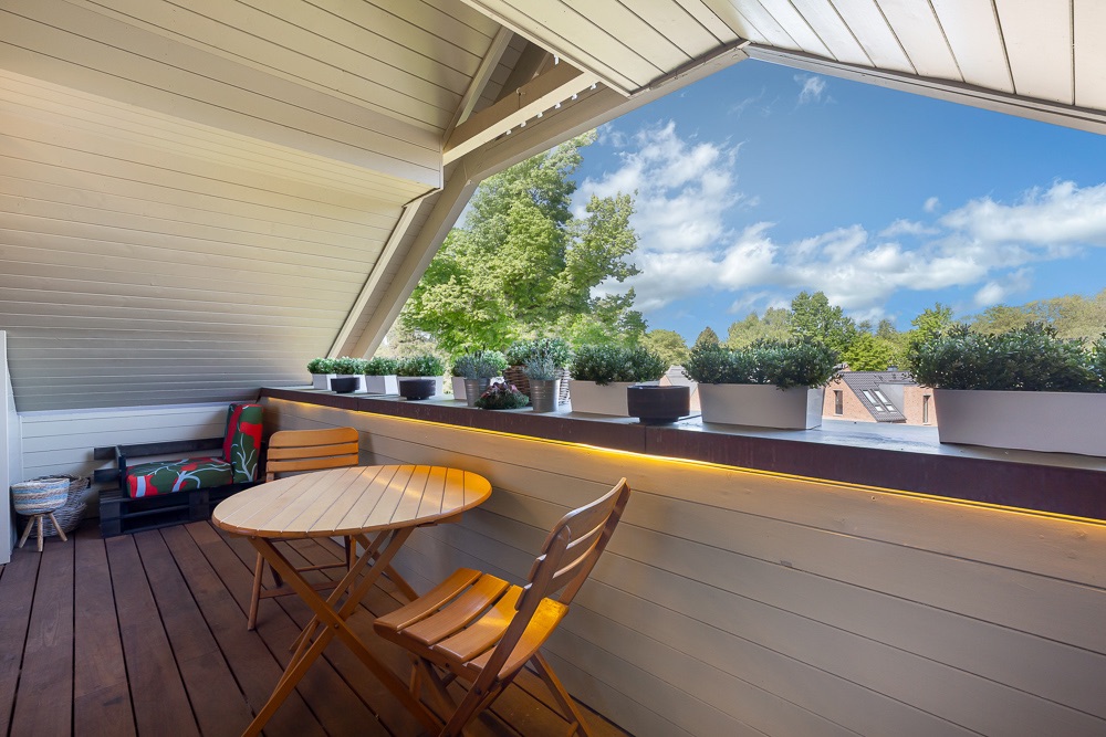Überdachter Balkon mit Holzboden, kleinem runden Tisch, zwei Stühlen und Pflanzkästen.