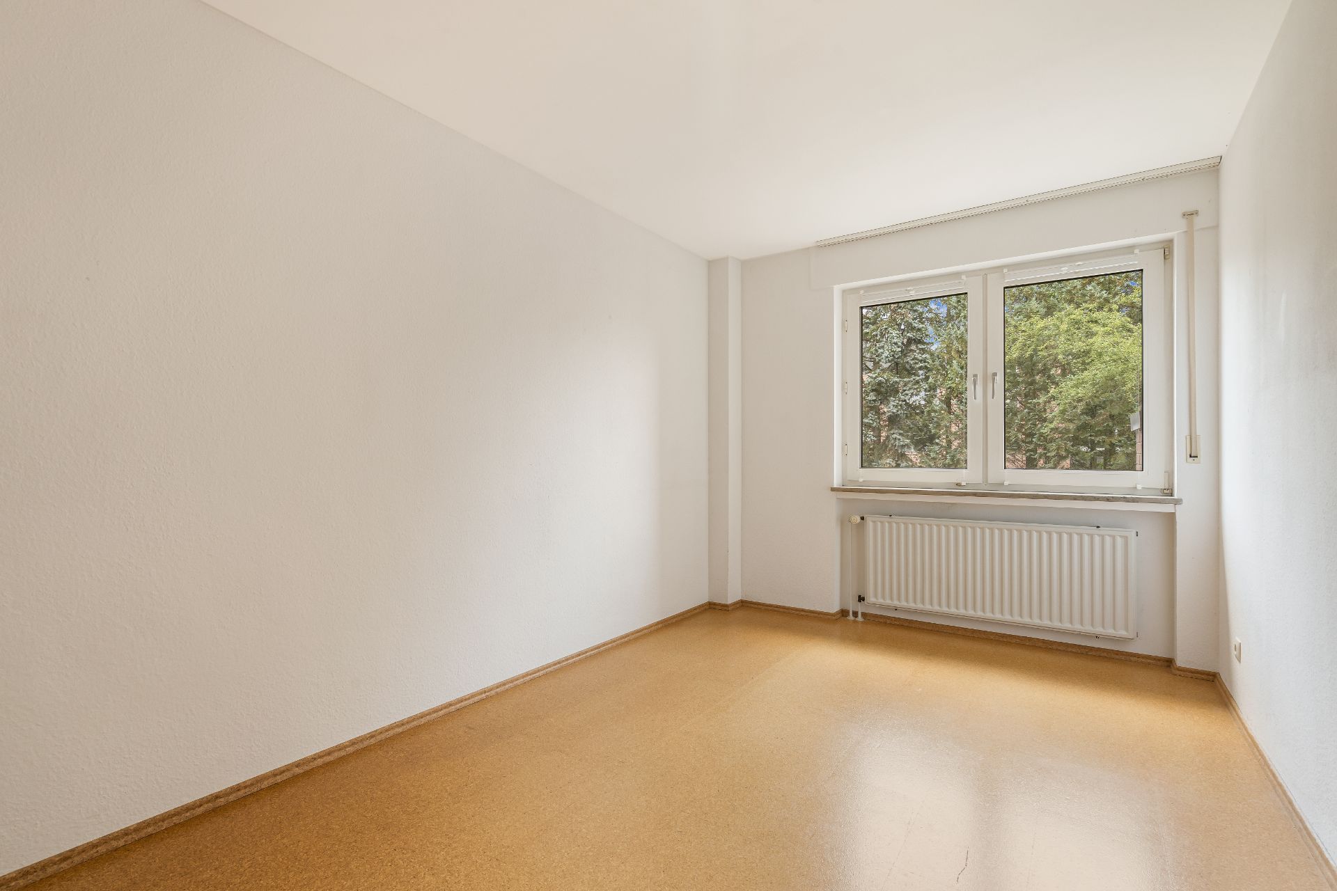 Leeres Zimmer mit hellem Boden, weißer Wand und großem Fenster mit Blick auf Bäume.