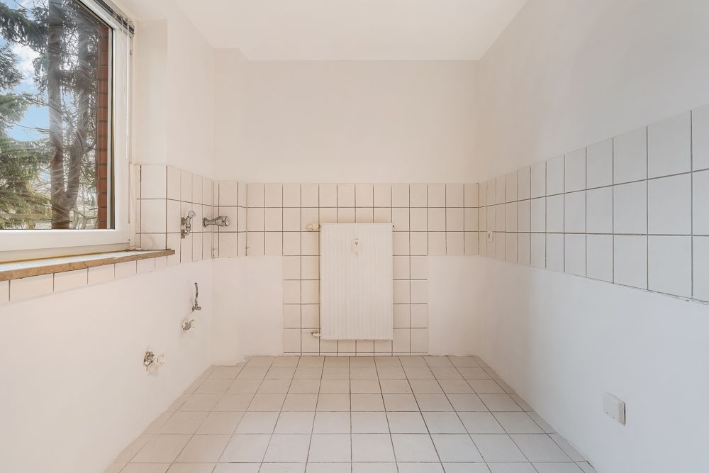 Leerer, gefliester Raum mit weißem Heizkörper und großem Fenster, Wasseranschlüsse sichtbar.