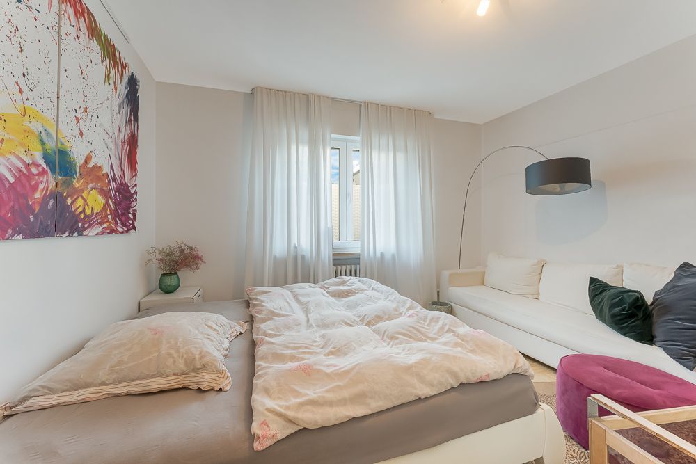 Helles Schlafzimmer mit Doppelbett, Sofa, großem Fenster und buntem Wandbild.