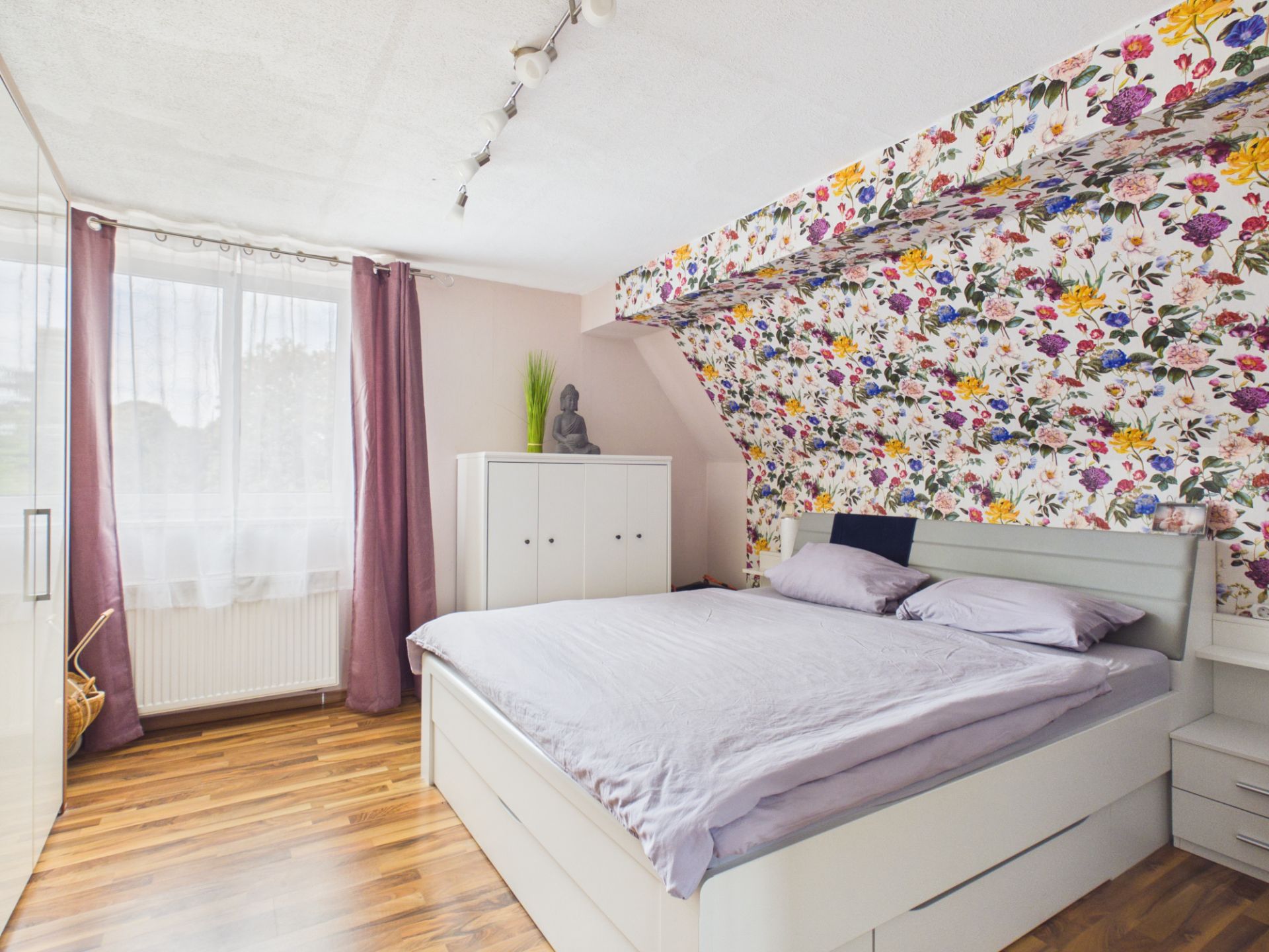 Helles Schlafzimmer mit Doppelbett, floral gemusterter Wand und großem Fenster mit lila Vorhängen.