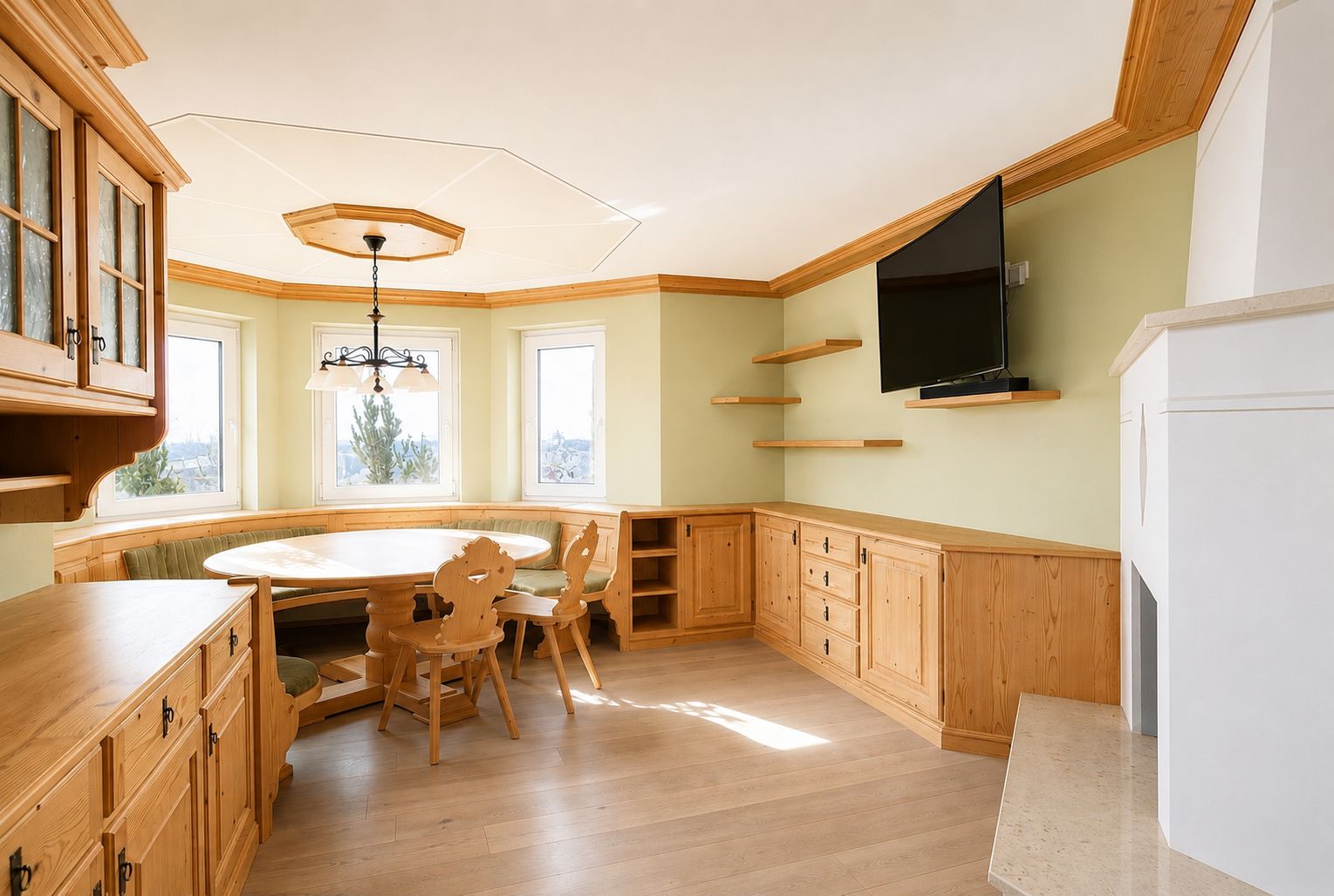 Heller Essbereich mit Eckbank, Holzmöbeln, runden Tisch, TV an der Wand und mehreren Fenstern.