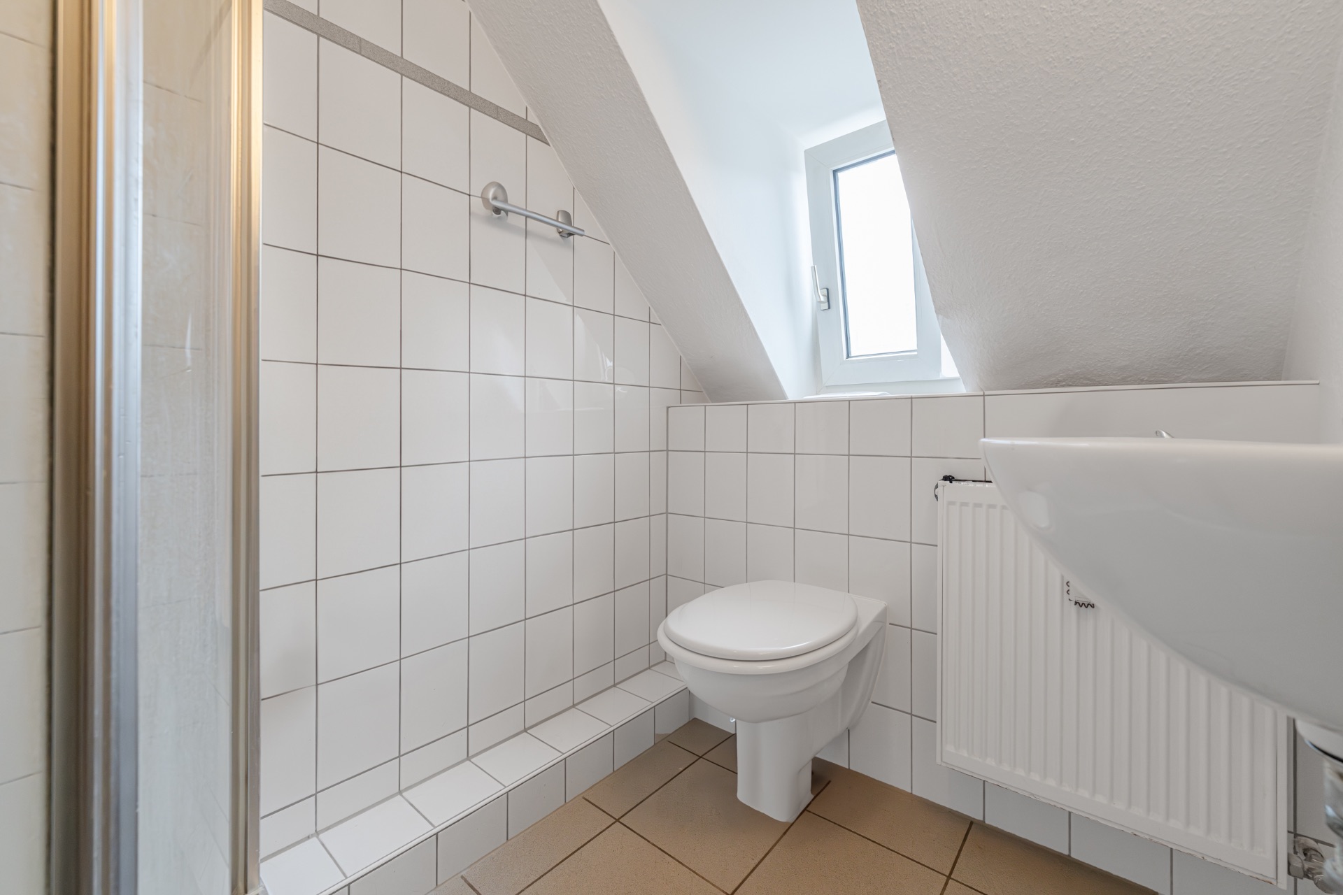 Modernes, weiß gefliestes Bad mit Hänge-WC, Waschbecken, Fenster und schräger Decke.