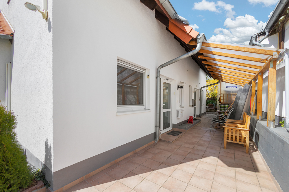 Überdachte Terrasse mit Fliesenboden, Holzbänken und weißen Wänden an einem Wohnhaus.
