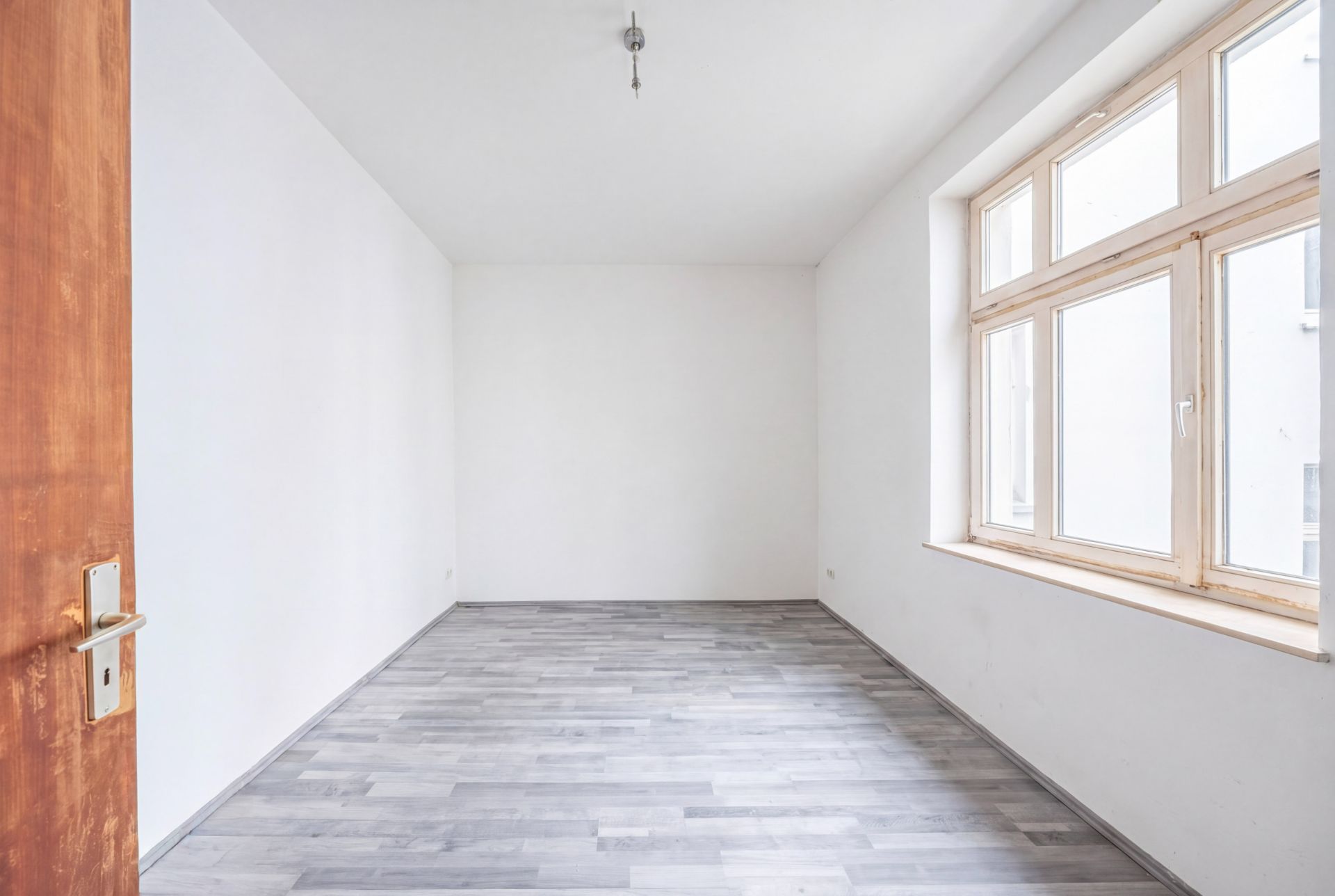 Leerer, heller Raum mit Holzfußboden und großem Fenster, weiße Wände.