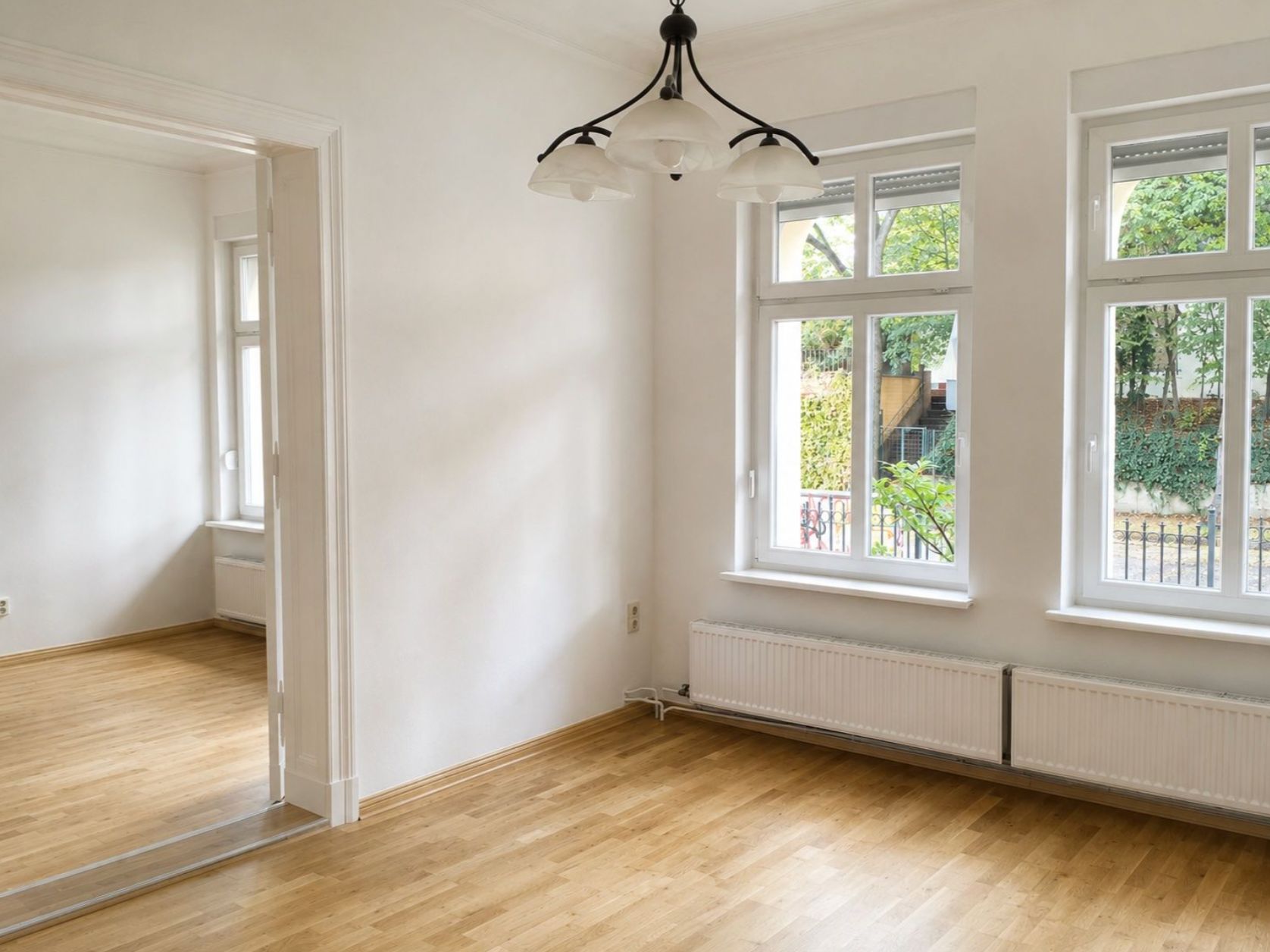 Helles Zimmer mit Holzfußboden, drei Fenstern, Heizkörper und Deckenlampe.