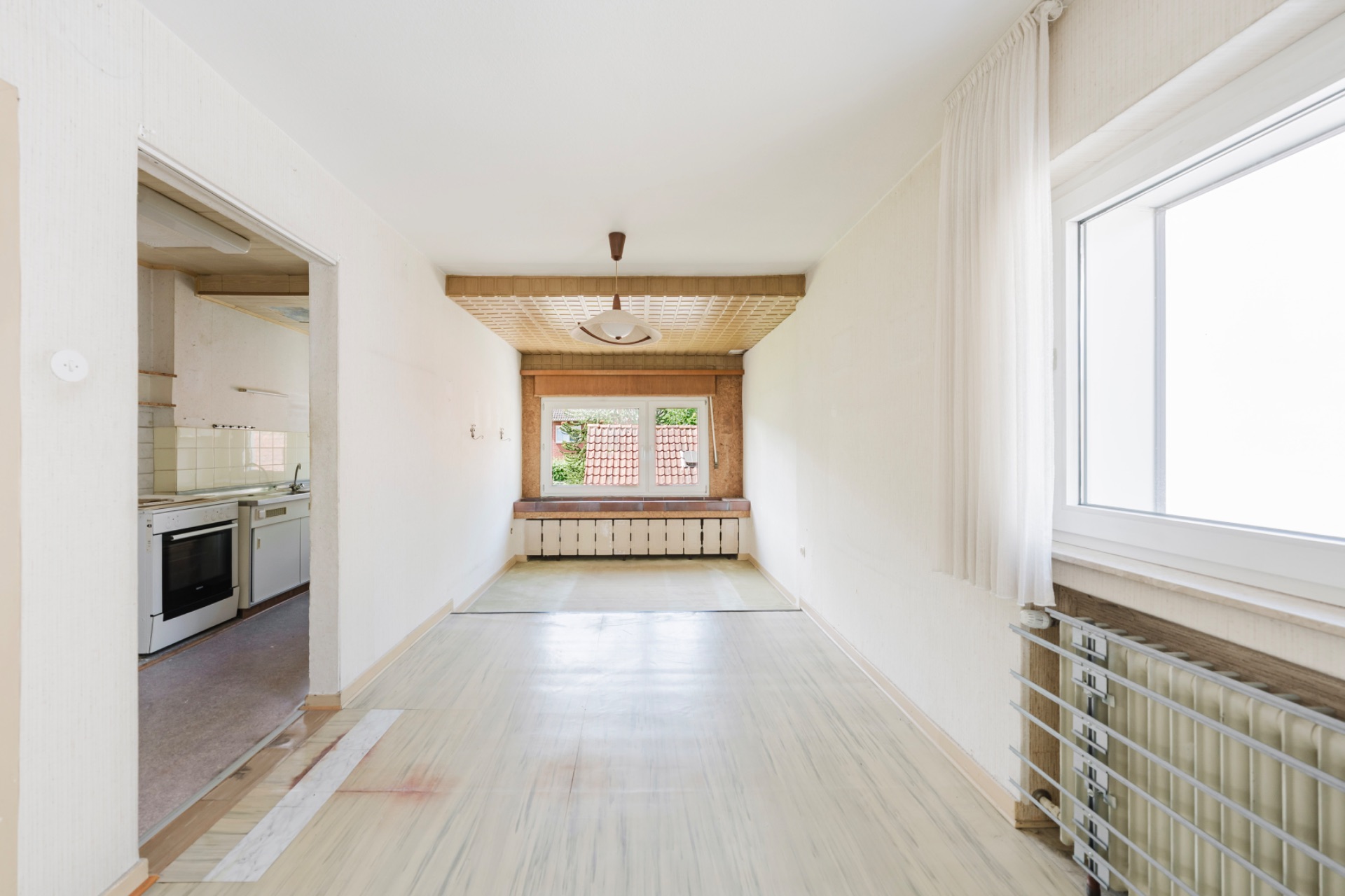 Heller, leerer Raum mit Fenster, Holzdecke am Ende und Zugang zur angrenzenden Küche.