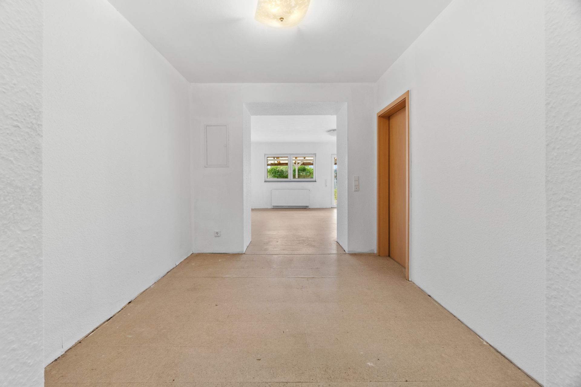 Heller Flur mit Blick in ein Wohnzimmer mit großen Fenstern und braunem Türrahmen rechts.