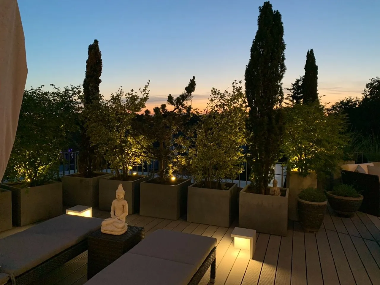 Beleuchtete Terrasse mit Pflanzen in großen Kübeln, Liegen und einer Buddha-Statue bei Sonnenuntergang.