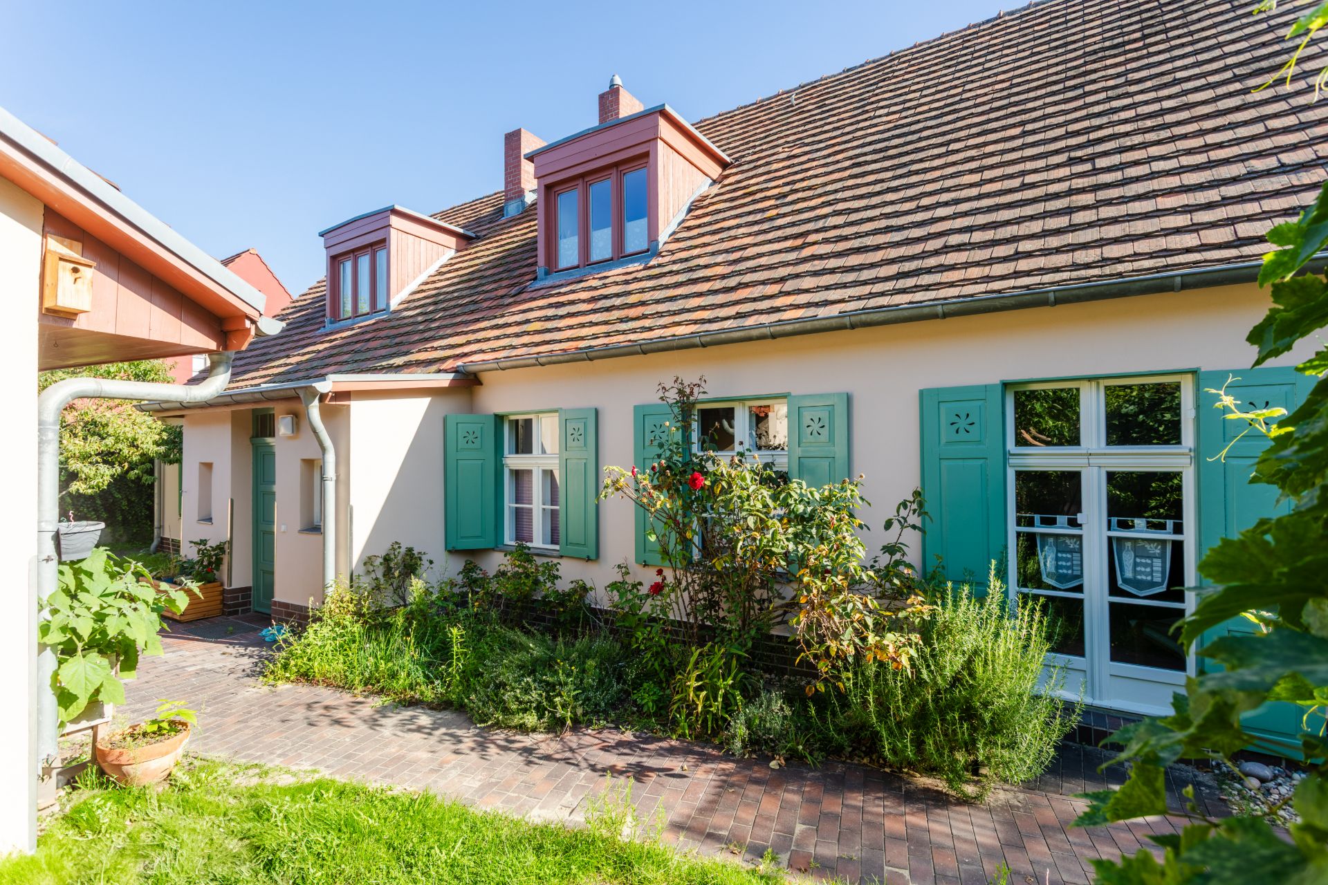 Wohnhaus mit grün gestrichenen Fensterläden und Garten im Vordergrund bei Sonnenschein.