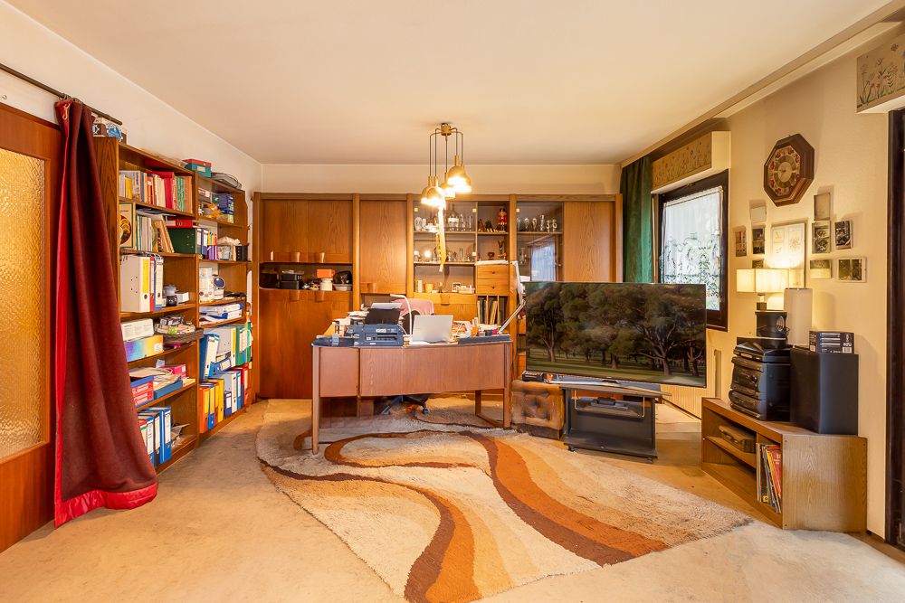 Wohnzimmer mit Schreibtisch, Bücherregalen, Teppich, Fernseher und Holzmöbeln.
