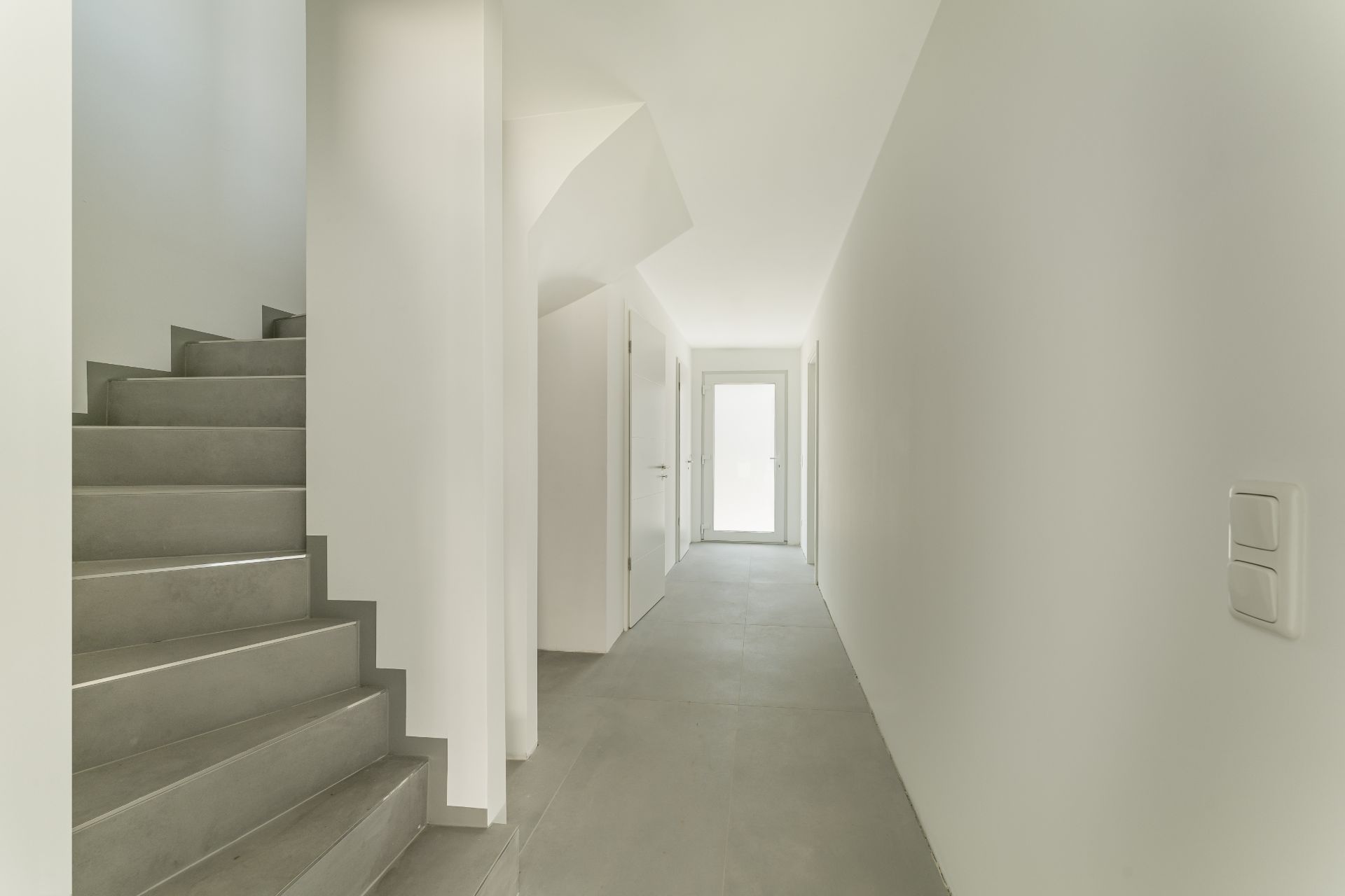 Heller Flur mit grauer Treppe auf der linken Seite und geschlossener Tür am Ende.