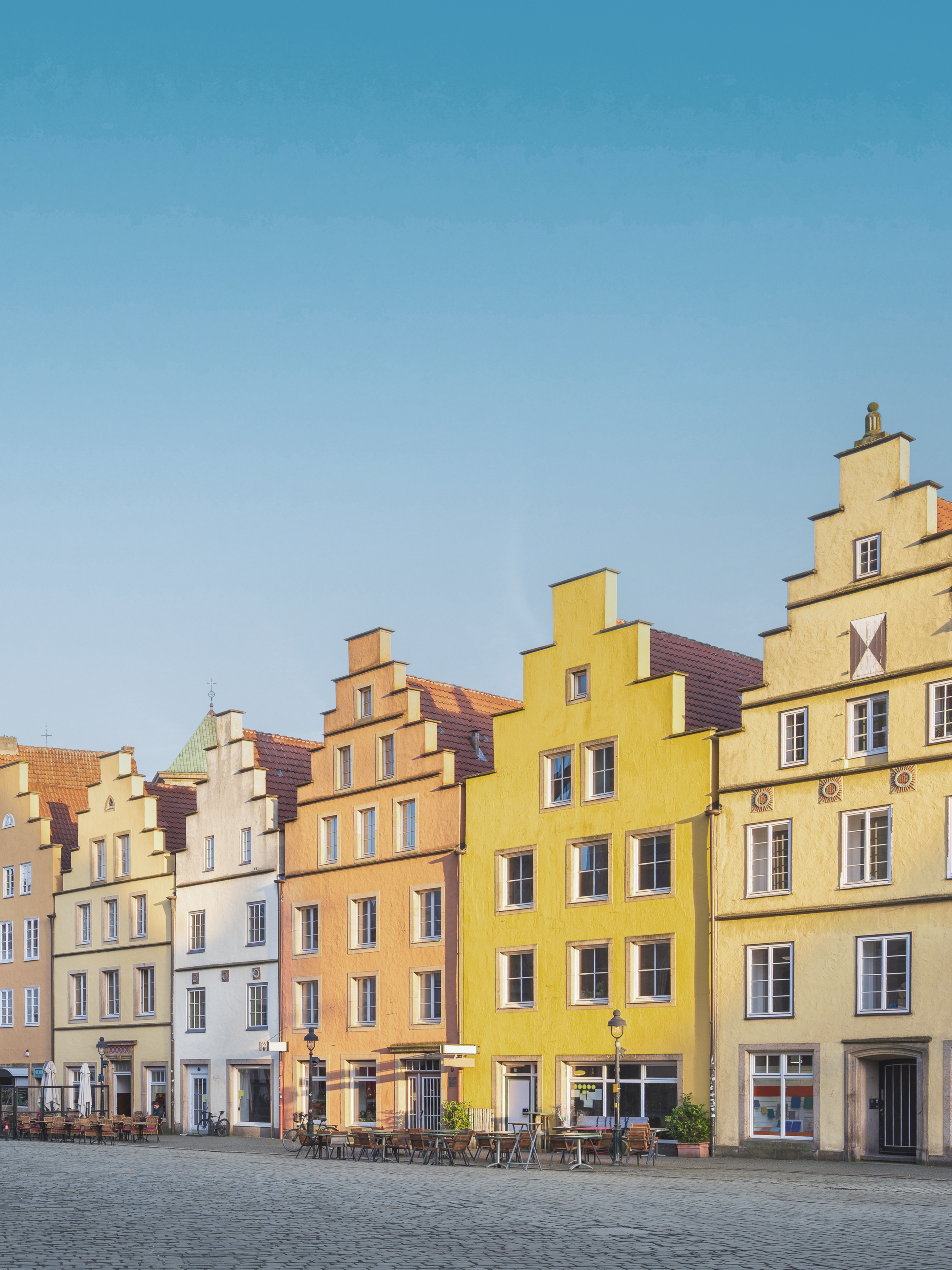 Mehrere bunte Wohnhäuser mit Stufengiebeln an einem Platz, davor stehen Tische und Stühle.