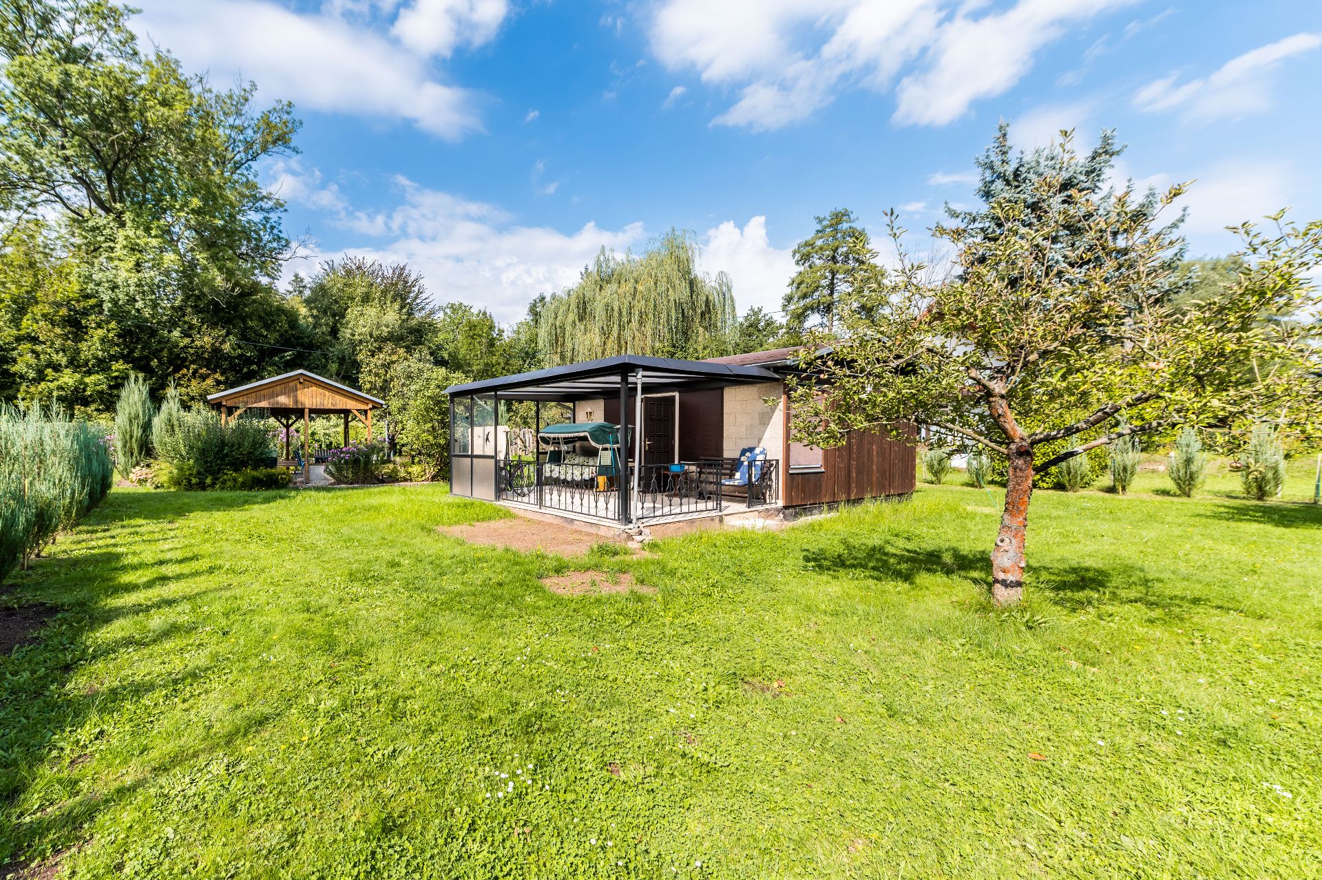 Kleines Haus mit überdachter Terrasse im großen, grünen Garten mit mehreren Bäumen.