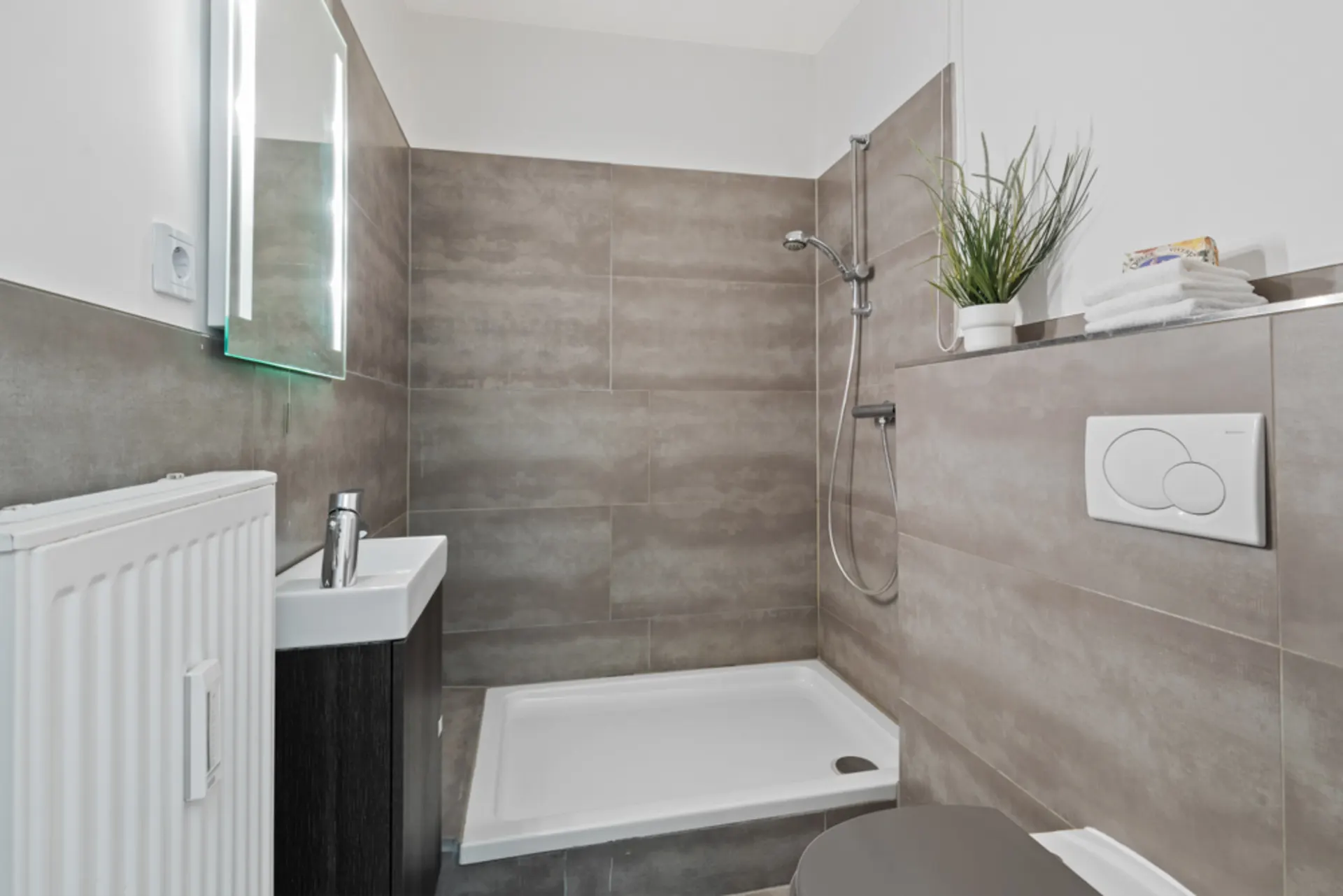 Modernes Bad mit Dusche, Waschbecken, Toilette und Wandspiegel, daneben steht eine Pflanze.