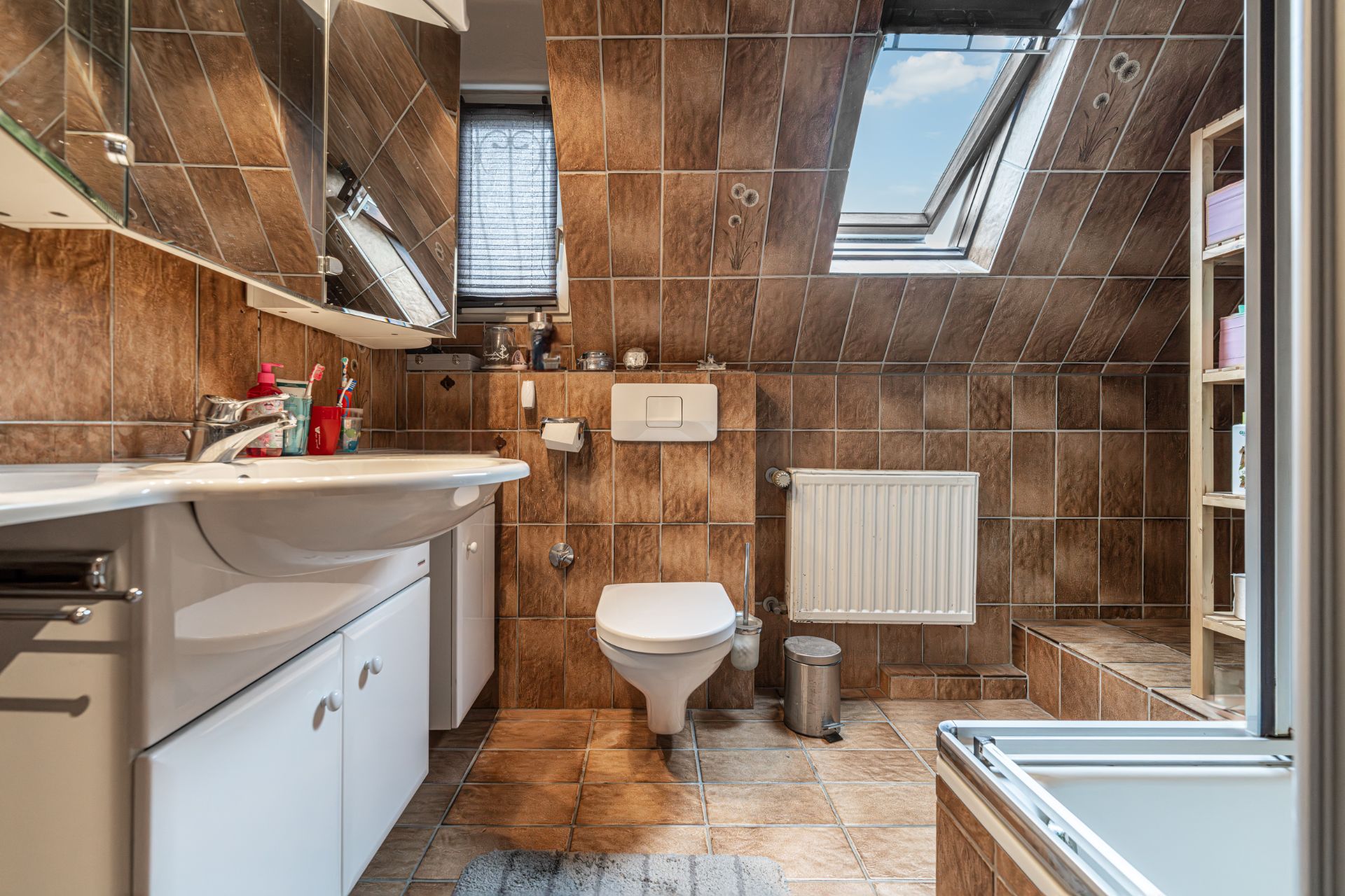 Badezimmer mit braunen Fliesen, Dachfenster, Waschbecken, WC und Regalen.