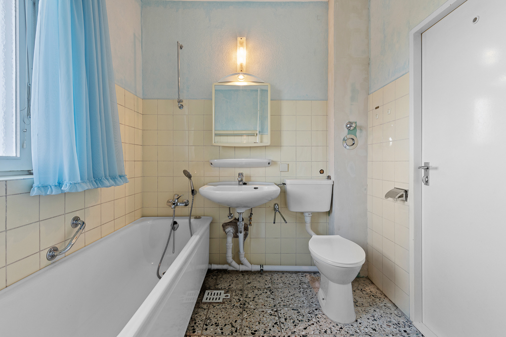 Helles Badezimmer mit Badewanne, Waschbecken, Spiegel, Toilette und blauem Duschvorhang.