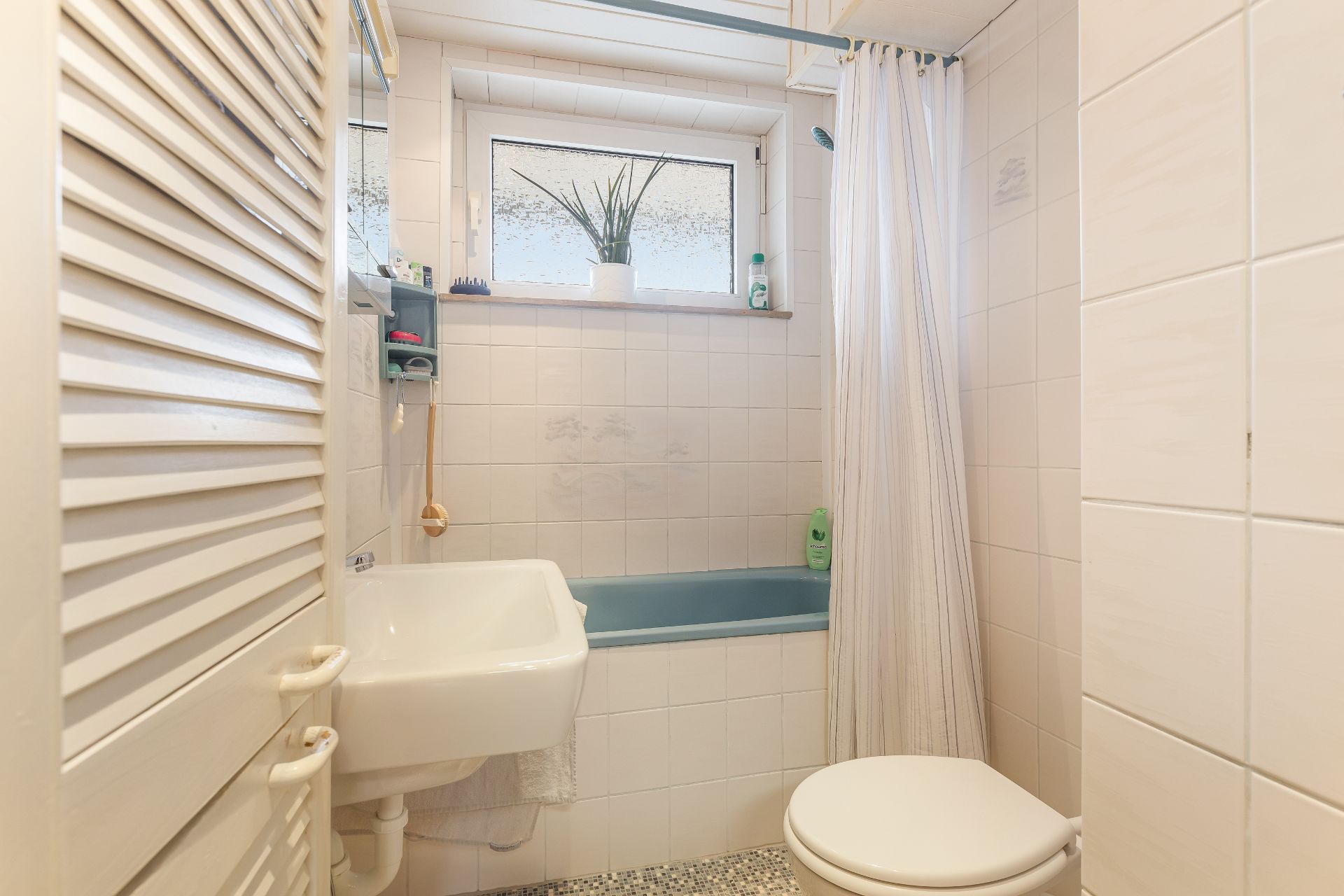 Kleines Badezimmer mit Badewanne, Waschbecken, Toilette und Fenster mit Pflanze.