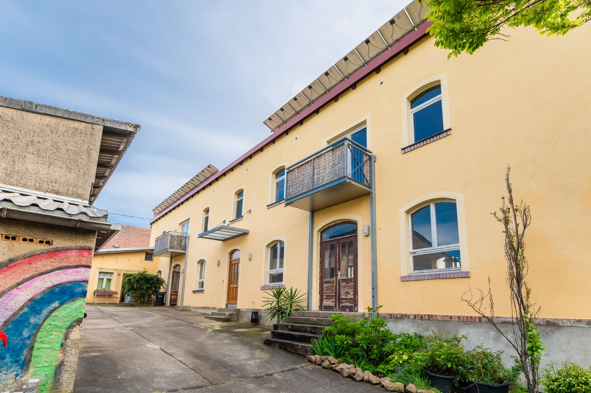 Gelbes Wohnhaus mit zwei Balkonen, großen Fenstern und kleinem bepflanztem Bereich im Innenhof.