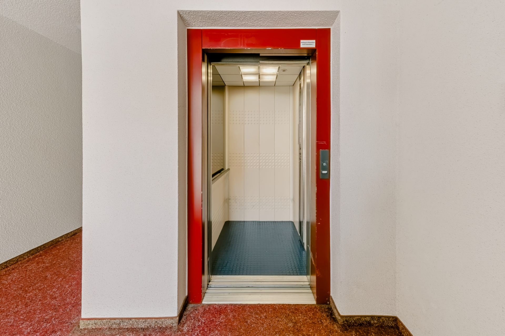 Offener Aufzug mit rotem Rahmen in einem Flur eines Wohngebäudes.
