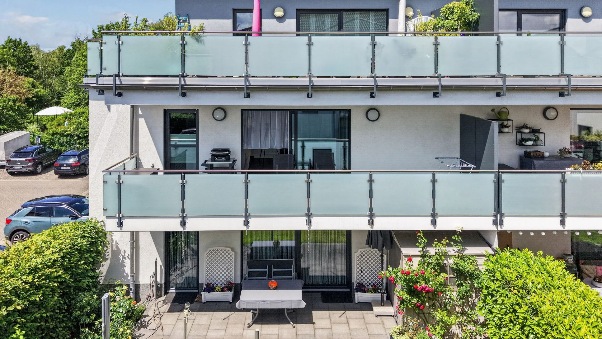 Zwei Balkone mit Glasgeländer an einem modernen Wohnhaus, darunter Terrasse mit Sitzbank und Pflanzen.